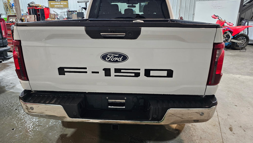 Rear view of a white Ford F-150 pickup truck tailgate in a garage setting, with the Ford logo and F-150 lettering on the tailgate.